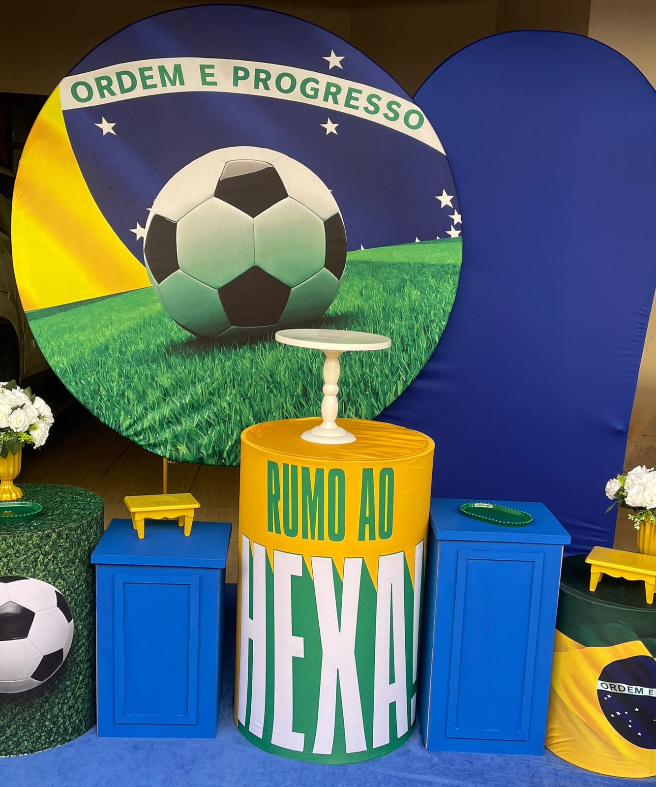 Decoração de festa com painel da bandeira do Brasil e bola de futebol, cilindros azuis, verde e amarelo