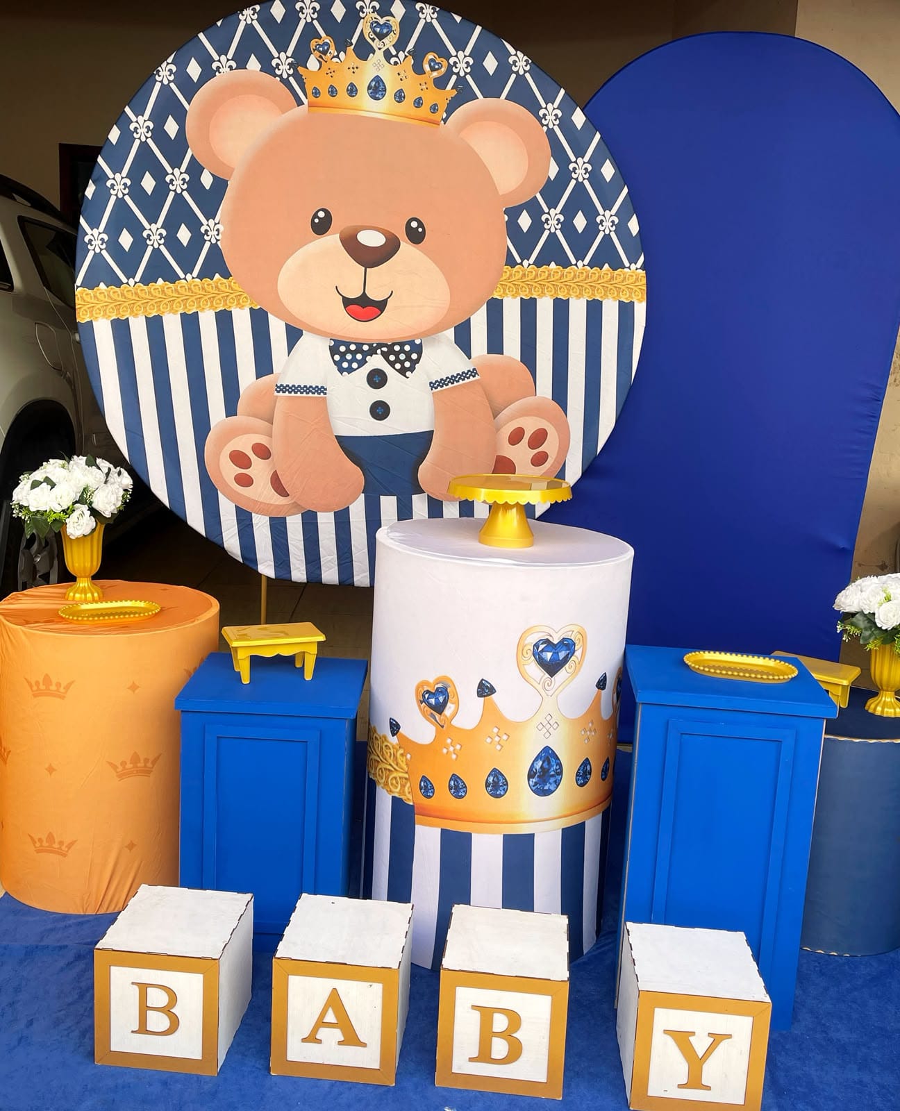 Decoração de festa com painel de ursinho príncipe com coroa em azul marinho, branco e dourado, cilindros azuis e cubos BABY